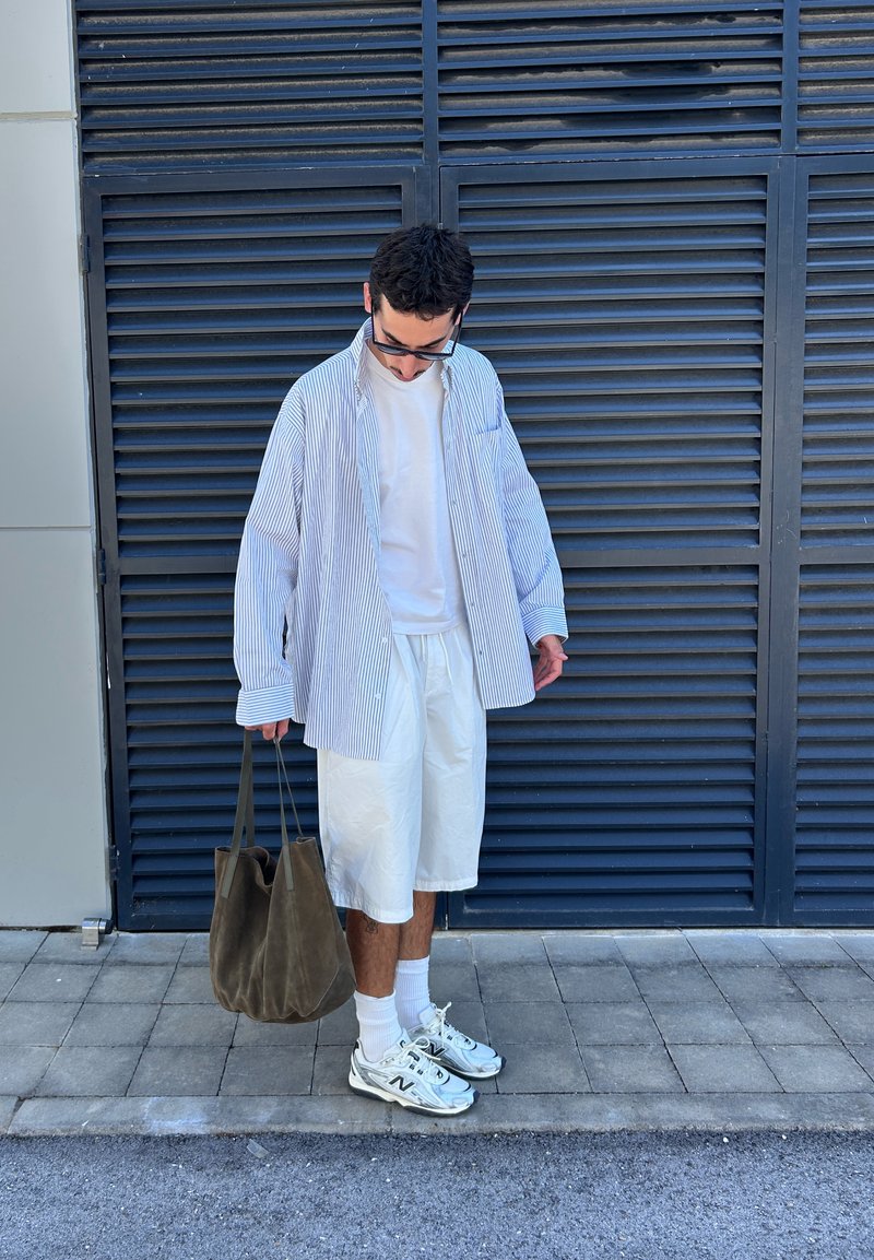 Homme portant une chemise rayée et un short blanc debout sur le trottoir, tenant un sac marron, regardant vers le bas devant des portes à volets bleu foncé.