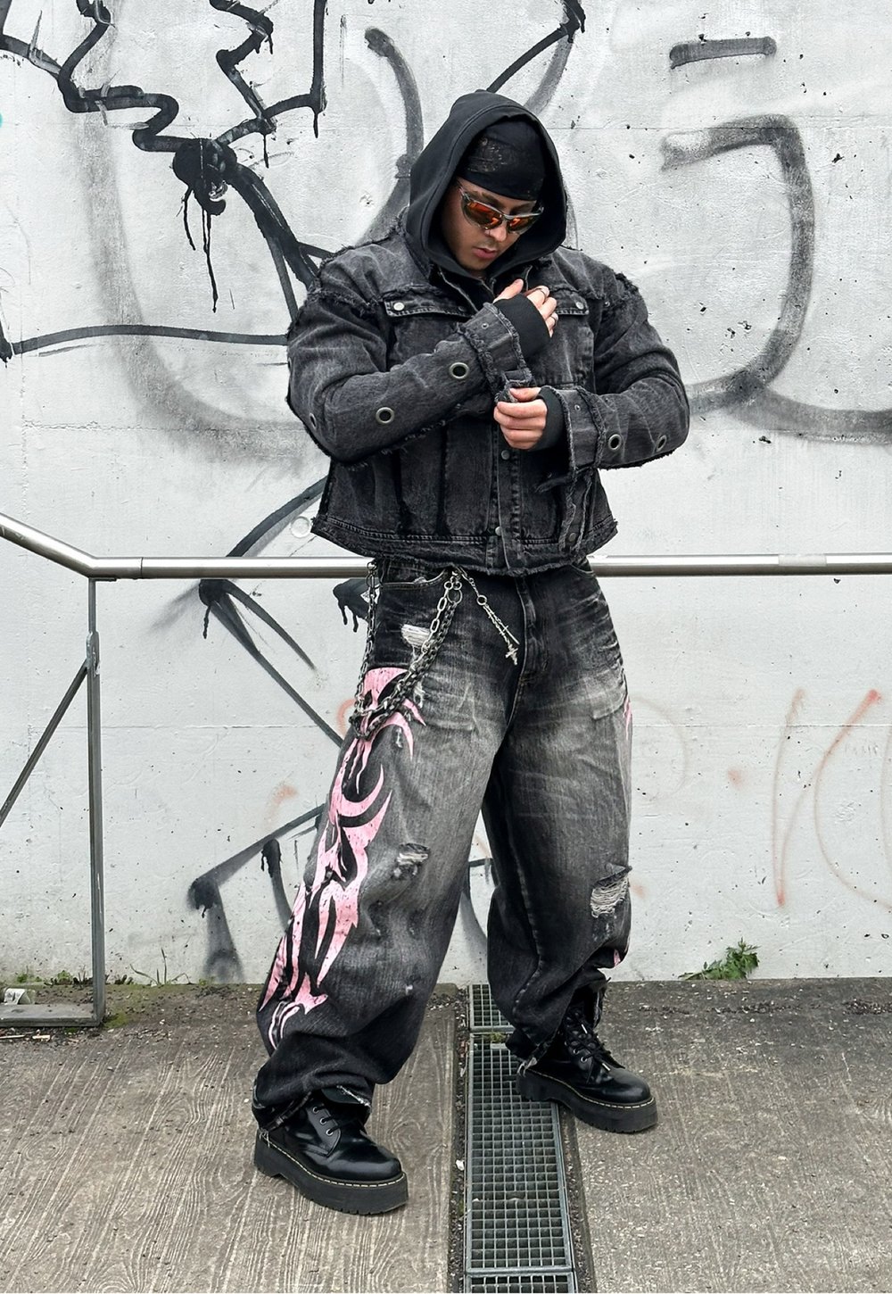 Μαύρο τζάκετ από τζιν με κοντή σχεδίαση και ασημένιες λεπτομέρειες, σε συνδυασμό με κατεστραμμένα μαύρα τζιν με ροζ φυλετικά μοτίβα. Ογκωτά μαύρα μποτίνια.