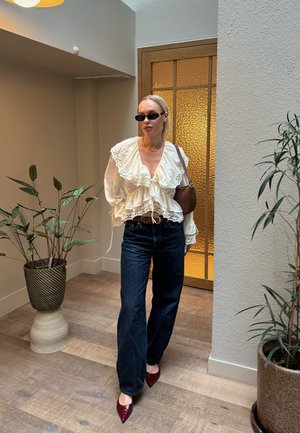 Mujer con blusa blanca con volantes, pantalones vaqueros anchos oscuros, zapatos puntiagudos burdeos, gafas de sol y bolso marrón de hombro, de pie en interiores junto a plantas.