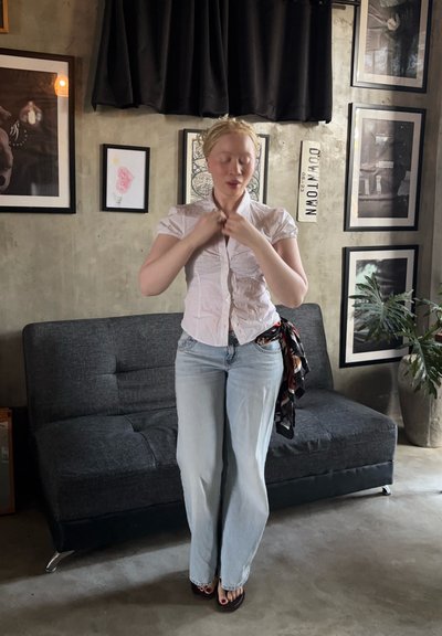Mujer con cabello claro, camisa blanca y vaqueros de pie en el salón, ajustándose el cuello, con un sofá oscuro y arte enmarcado en la pared detrás de ella.