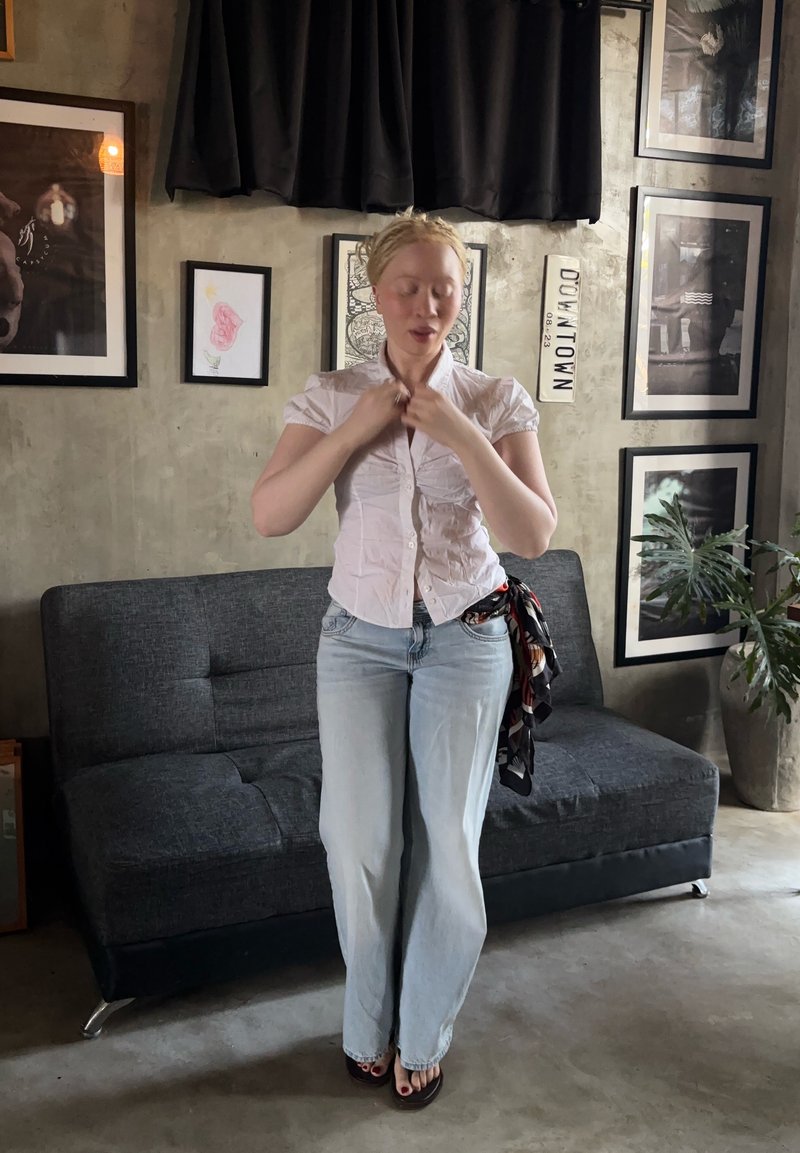 Femme aux cheveux clairs portant une chemise blanche et un jean, debout dans le salon, ajustant son col, avec un canapé sombre et une œuvre encadrée sur le mur derrière elle.
