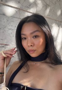 Primer plano de una persona con cabello largo y oscuro, vistiendo una blusa negra y una cinta oscura alrededor del cuello. Los accesorios notables incluyen un collar delicado y un bolso.