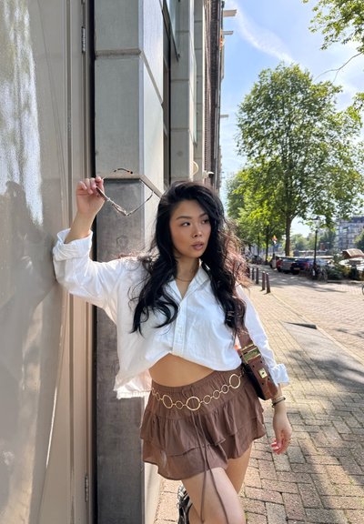 Una camisa blanca recortada de botones, una falda marrón con capas y acentos circulares dorados, y un bolso de hombro contra un fondo de ciudad.