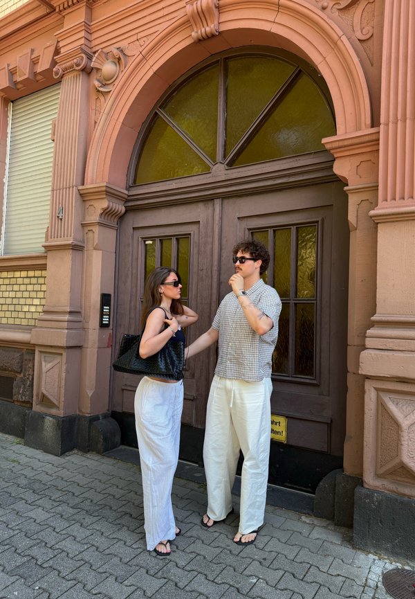 A man and woman wearing sunglasses stand talking in front of a large decorative brown door on a cobblestone sidewalk.