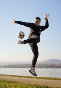 Eine Person, die ein schwarzes Langarmshirt und dunkle Hosen trägt, tritt gegen einen mehrfarbigen Fußball, während sie auf Gras neben einem Weg steht.