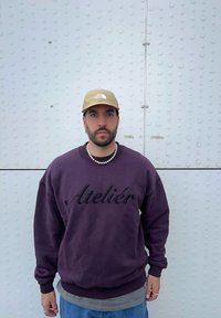 Sweatshirt violet avec "Atelier" en lettres noires, superposé à une chemise grise, associé à un jean bleu et une casquette beige.