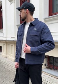 Veste en coton bleu marine avec deux poches poitrine et fermeture à boutons. Portée sur une chemise blanche, associée à un pantalon noir et une casquette.