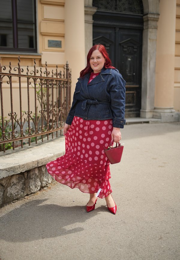 Femme aux cheveux roux portant une robe rouge à pois, une veste en jean, des chaussures rouges et tenant un sac à main rouge, debout près d'une clôture en fer forgé.