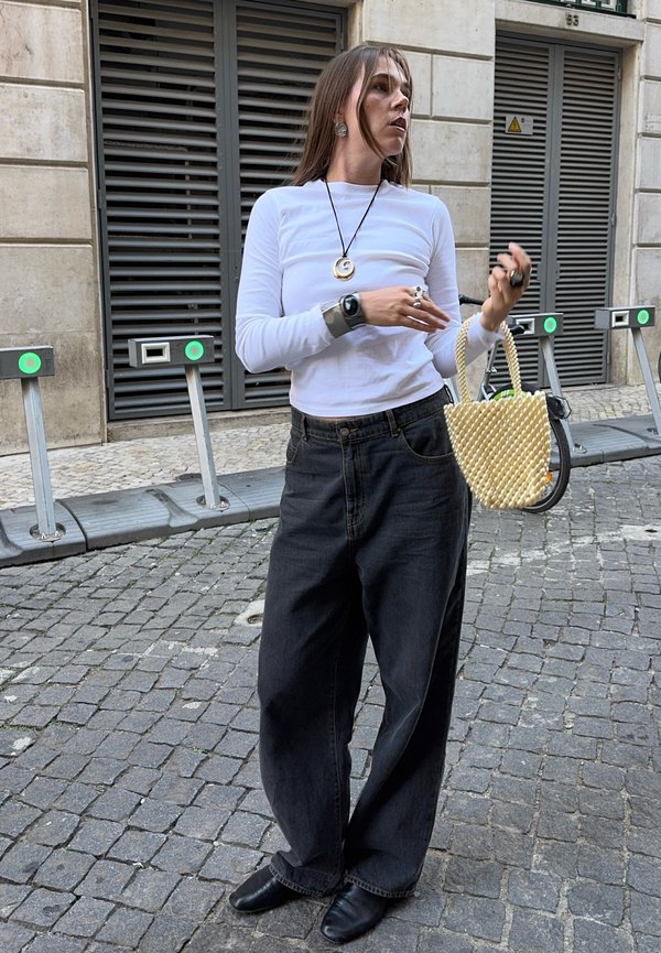 Femme en chemise blanche à manches longues et pantalon large noir se tient dans une rue pavée, tenant un sac à main orné de perles et portant plusieurs bagues.