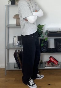 Inimene kannab halli kihilise pluusi, musti laiaid pükse ja musti sportlikke jalatseid, seistes käed risti metallriiuli kõrval, millel on kõlarid ja taimed.