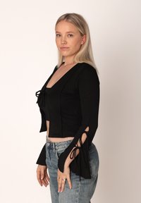 Μαύρο μακρυμάνικο crop top με V-λαιμόκοψη και λεπτομέρειες δεσίματος στις μανσέτες, συνδυασμένο με ανοιχτό μπλε ψηλόμεσο τζιν. Λείος υφιστάμενος.