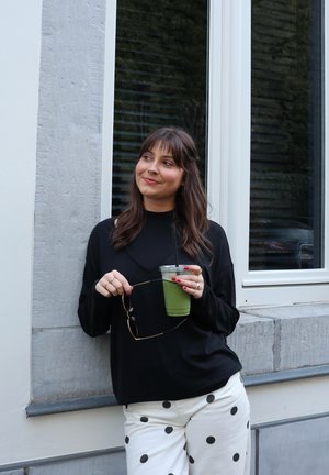 Joven mujer con camiseta negra y pantalones blancos con lunares se apoya en una pared, sosteniendo gafas de sol y una bebida verde con hielo y pajilla.