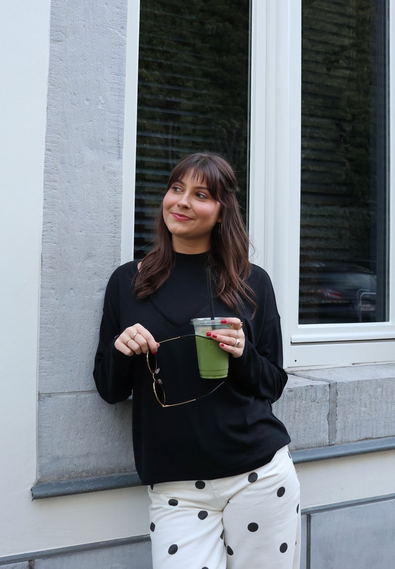 Jeune femme en haut noir et pantalon blanc à pois s'appuyant contre un mur, tenant des lunettes de soleil et une boisson glacée verte avec une paille.
