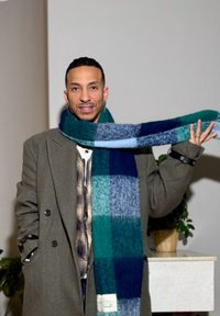 Longue écharpe à carreaux en bleu sarcelle, bleu foncé et bleu clair avec une texture tissée. Portée drapée sur les épaules, associée à un manteau vert olive.