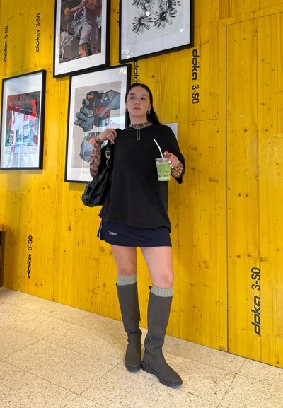 Camisa negra de gran tamaño, falda corta azul marino, botas gris claro hasta la rodilla con puños acanalados. Sosteniendo una bebida verde, de pie contra una pared amarilla de madera.