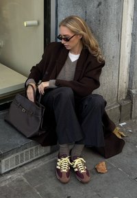 Femme assise sur un trottoir portant un manteau foncé, un pantalon à fines rayures, des baskets bordeaux, un pull marron clair, des lunettes de soleil, et tenant un grand sac à main foncé.