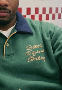 Grön fleecetröja med polokrage i marinblått. Har gyllene broderad text "Bllvts Original Clothing" på bröstet och en knappslå.