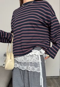 Personne portant un haut à manches longues rayé bleu marine, pantalon gris avec bordure en dentelle blanche et un nœud, tenant un petit sac à main beige avec un détail en perles.