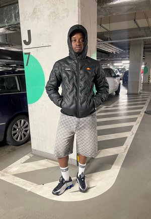 Chaqueta de plumas negra con capucha, pantalones cortos grises con estampado y zapatillas negras y grises. De pie en el suelo de un garaje con vehículos de fondo.