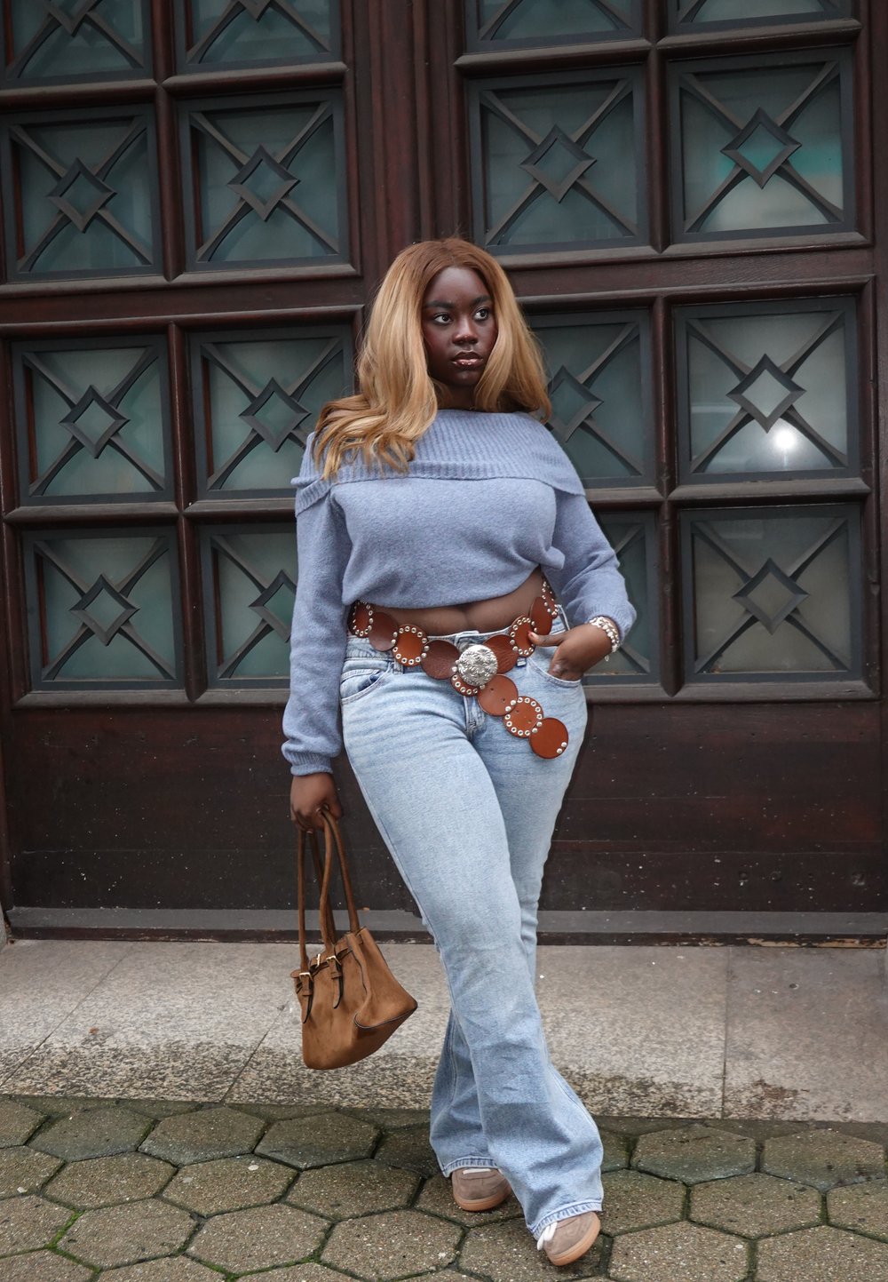 Lys blå kort genser, høytlivs lyse jeans, brunt skinnbelte med sirkulære detaljer, tanfarget håndveske og beige sko. Sekskantet brosteinsbakgrunn.