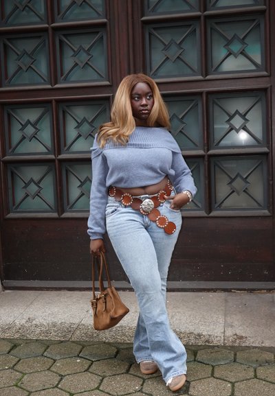 Suéter corto azul claro, jeans de tiro alto claros, cinturón de cuero marrón con detalles circulares, bolso marrón claro y zapatos beige. Fondo de pavimento hexagonal.