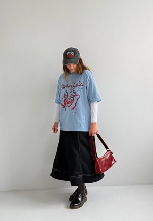 Persona joven con gorra negra, camiseta gráfica azul clara sobre mangas largas blancas, falda midi negra, zapatos negros, sosteniendo un bolso rojo.