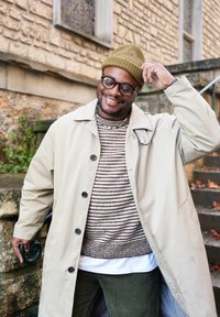 Beige long coat over a brown striped jumper, dark green corduroy trousers, and a green knitted beanie. Eyeglasses and textured materials feature prominently.