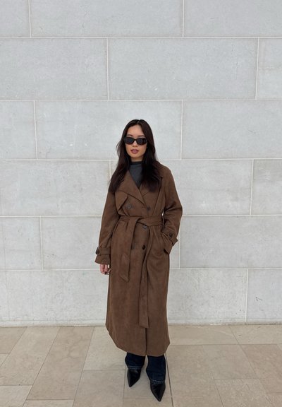 Mujer con cabello largo y oscuro, usa gafas de sol negras, un abrigo largo marrón con cinturón, pantalones negros y zapatos negros puntiagudos, de pie contra una pared de piedra.