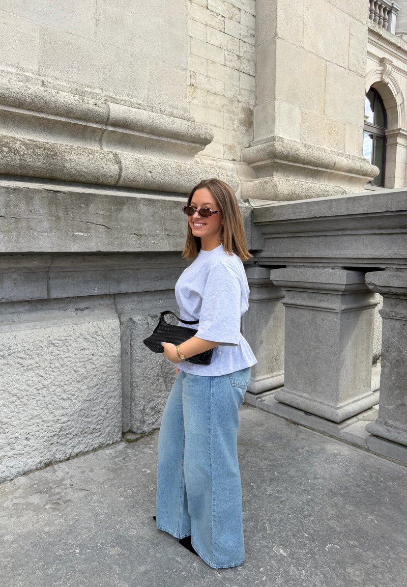 Femme portant des lunettes de soleil, une chemise blanche et un jean bleu large tenant un sac noir, debout près de grandes colonnes en pierre sur un trottoir gris.