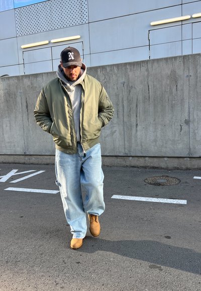 Chaqueta bomber verde sobre una sudadera gris, jeans sueltos de color azul claro y botas marrones. Una gorra negra con un logo blanco completa el conjunto.