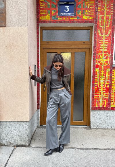 Chaqueta de cuero marrón con cuello acanalado, top corto gris, pantalones de pierna ancha a rayas grises y botas negras de punta afilada frente a una pared de mosaico colorido.
