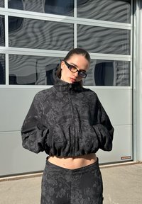 Casaco preto cortado com textura floral, gola alta e punhos elásticos, combinado com leggings de cintura alta coordenadas.