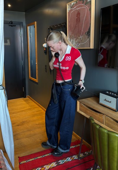Joven mujer con top rojo y vaqueros anchos usando un teléfono de disco negro en una habitación moderna con suelo de madera y paredes oscuras.