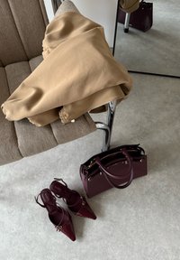 Veste beige sur l'accoudoir d'un canapé beige, assortie à des escarpins bordeaux à bouts pointus et un sac à main assorti sur un sol gris clair.