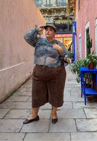 Bluză din dantelă albastră transparentă cu detalii cu volane, culote din bumbac corduroy maro și pantofi negri. Ținând o geantă mică, cu un fundal de plante în ghiveci și lumini de tip fairy.