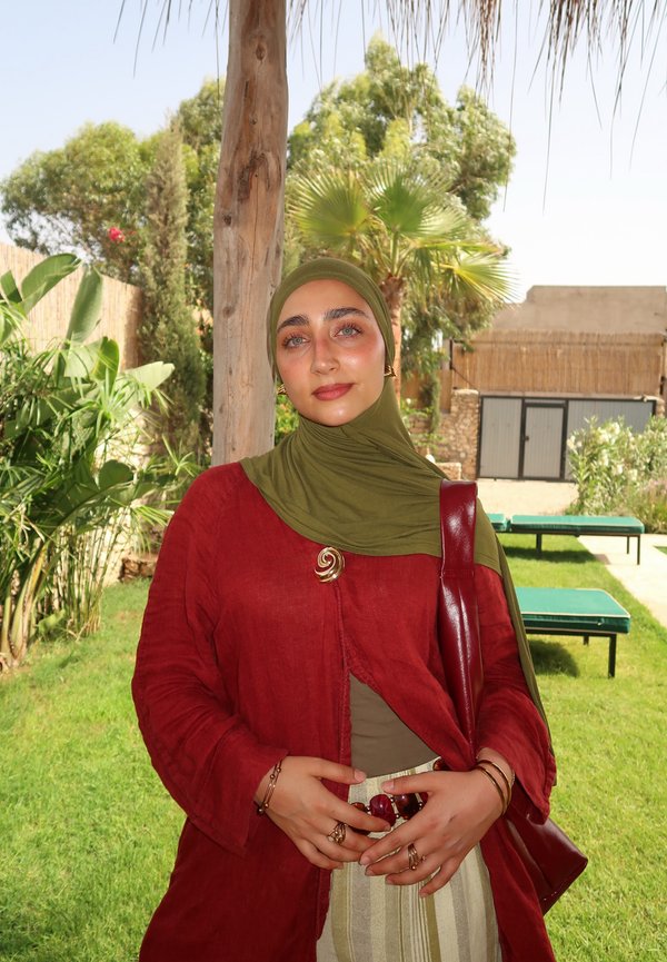 Femme portant un hijab vert olive et une veste rouge, debout à l'extérieur près d'un poteau en bois, avec des plantes vertes et des chaises longues en arrière-plan.