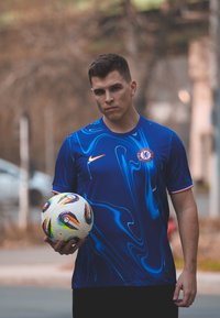 Blue athletic jersey featuring abstract swirl patterns, a Nike logo, and the Chelsea emblem. The person is holding a white soccer ball with colourful accents.