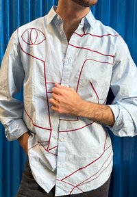 Lyseblå skjorte med knapper, preget av røde strektegninger på forsiden. Stoffet ser ut til å være teksturert, med en avslappet passform og oppbrettede ermer.