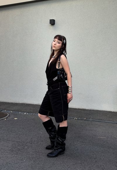 Top negro sin mangas con diseño drapeado, pantalones cortos de mezclilla negros, botas altas negras con caída hasta la rodilla y bolso negro con acentos plateados.