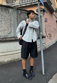 Šviesiai mėlynas tekstūruotas marškinėlių švarkelis, juodos šortai, juodos kojinės ir juodi loaferiai. Juodas krepšys pakabintas ant vienos peties.