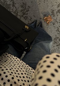 Άτομο που φοράει μαύρα μυτερά παπούτσια και μπλε τζιν, κρατώντας μια μαύρη τσάντα, στέκεται σε άσφαλτο με ένα ξερό φύλλο κοντά.