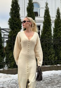 Femme avec une queue de cheval blonde portant une robe en tricot crème boutonnée et des lunettes de soleil, tenant un sac à main noir, debout près d'une clôture en fer et de grands arbres persistants.