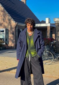 Ναυτικό παλτό, πράσινο πλεκτό ζακέτα, μαύρο παντελόνι wide-leg, στρωμένες αλυσίδες, ακουστικά, όρθια σε εξωτερικό χώρο μπροστά σε ένα τούβλινο σπίτι.