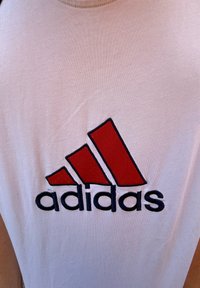 Vit ärmlös skjorta med en stor röd och svart Adidas-logotyp. Tyget verkar mjukt med en slät textur.