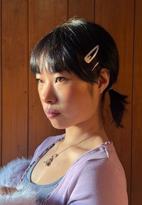 Femme aux cheveux noirs attachés en queue de cheval avec des barrettes, regardant intensément sur le côté, portant un haut violet clair et un collier doré, devant un fond de panneaux en bois.