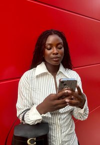 Camicia bianca a righe con un ampio colletto, che tiene uno smartphone con una custodia strutturata, abbinata a una borsa nera davanti a un muro rosso.