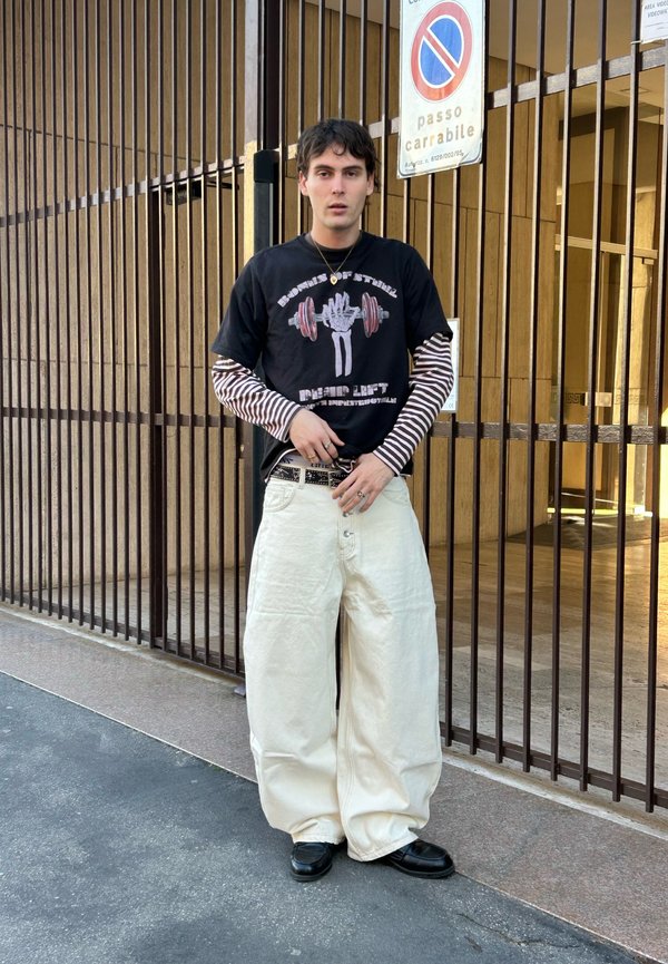 Tânăr purtând un tricou cu dungi pe mânecă lungă sub un tricou cu imprimeu, pantaloni largi albi și pantofi negri, stând lângă o poartă metalică cu un semn de staționare interzisă.