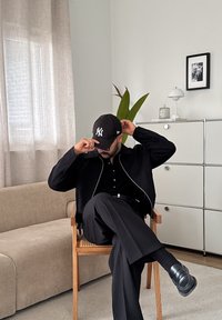 Mann i svart antrekk og en New York Yankees caps som sitter med beina krysset på en trestol i et moderne stue.