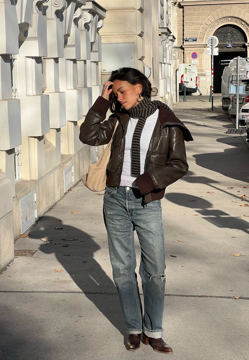 Donna con giacca di pelle marrone, sciarpa a righe, jeans e stivali, in piedi su un marciapiede soleggiato di una città accanto a una facciata di edificio ornata.