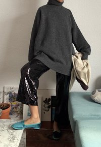 Personne portant un pull à col roulé oversize gris foncé, un pantalon noir à sequins et des chaussures slip-on en velours bleu, tenant une veste beige clair.